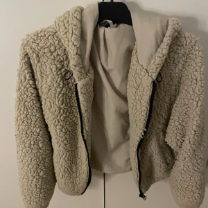 Beige teddyjacka med huva - Mysig beige teddyjacka med huva och dragkedja framtill. Jackan har en fluffig, fårullsliknande yta och är perfekt för kyliga dagar. Modellen är croppad och har en avslappnad passform som ger en cool och trendig look.