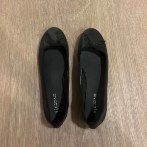 Svarta ballerinaskor i storlek 39 - Klassiska svarta ballerinaskor från Divided by H&M i storlek 39. Skorna har rund tå, liten rosett framtill och är tillverkade i syntetmaterial. Perfekta för en clean och enkel look.