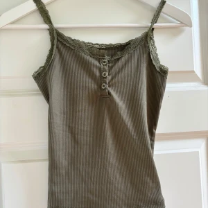 Olivgrön ribbad linnetopp från Dazy - Supersöt olivgrön ribbad linnetopp från Dazy med spetskant och smala axelband. Toppen har tre knappar framtill som detalj och är i mjuk, stretchig bomullsmix. Perfekt att styla med jeans eller kjol för en chill look.
