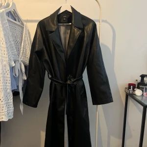 Svart lång skinn trenchcoat Boohoo - Säljer en svart lång trenchcoat i skinnimitation från Boohoo. Jackan har klassisk krage, bälte i midjan och raka ärmar. Passformen är loose och passar också 38 