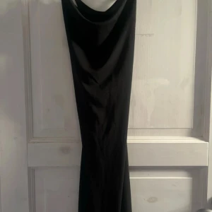 Svart långklänning med draperad hals - Snygg svart långklänning med draperad halsringning och figurnära passform. Klänningen är ärmlös och har en stilren, minimalistisk look som passar perfekt till en cool och elegant outfit. Materialet känns mjukt och följsamt mot kroppen.