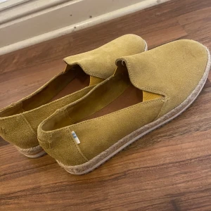 Gula mocka loafers från TOMS - Snygga loafers från TOMS i gul mocka med rund tå och platt sula. Skorna har en repdetalj runt sulan och elastiska partier vid insteget för extra komfort. Perfekt för dig som gillar en chill och stilren look med lite boho-vibe.