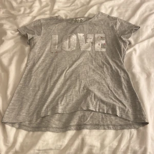 Grå t-shirt med paljetter från KappAhl - Snygg grå t-shirt från KappAhl med korta ärmar och rund hals. Framsidan har stora bokstäver i rosa paljetter som stavar 'LOVE'. Mjuk och skön bomullskvalitet, perfekt för dig som gillar lite extra bling i vardagen.