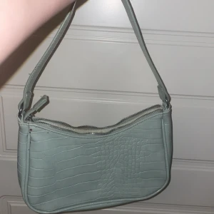 Ljusgrön axelväska från WOW - Snygg ljusgrön axelväska från WOW med croco-mönstrad yta i syntetmaterial. Väskan har en bred axelrem och praktisk dragkedja upptill. Perfekt storlek för det viktigaste och ger en cool vibe till din outfit.