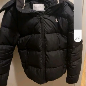 Svart pufferjacka från Calvin Klein - Snygg svart pufferjacka från Calvin Klein Jeans med klassisk CK-logga på bröstet och ärmen. Jackan har huva, dragkedja framtill och är vadderad för extra värme. Perfekt för kalla dagar och riktigt stilren look.
