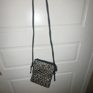 Leopardmönstrad axelväska från Glitter - Snygg liten axelväska med leopardmönster från Glitter. Väskan har svart axelrem, dragkedja upptill och en brun insida. Perfekt storlek för mobilen och småsaker. Materialet är syntet med glansig finish och detaljer i guld.