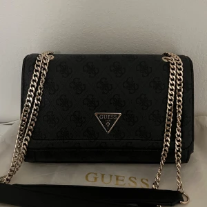 Svart axelväska från Guess med kedja - Snygg svart axelväska från Guess med diskret mönster och guldfärgad kedja som axelrem. Väskan har ett klassiskt trekantsformat Guess-emblem framtill och flera fack inuti för smart organisering. Materialet är slitstark syntet med lyxig känsla. OBS väskan är original och aldrig använd.