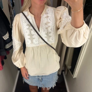 Beige puffig blus - Säljer en beige blus med broderade vita detaljer framtill och på ärmarna. Blusen har puffiga långärmar med resår vid ärmslut och en luftig passform. Har verkligen aldrig använt den så super nyskick! Nypris 699kr