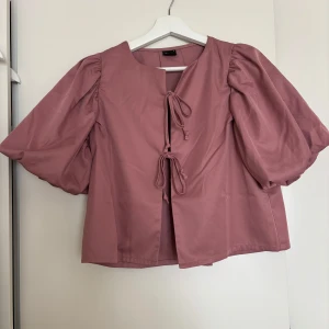 Rosa blus med puffärm från Gina Tricot - Supersöt rosa blus från Gina Tricot med puffiga korta ärmar och två knytningar framtill. Blusen har en rak passform och är tillverkad i ett slätt tyg som känns mjukt mot huden. Perfekt för dig som gillar trendiga detaljer och en feminin look.