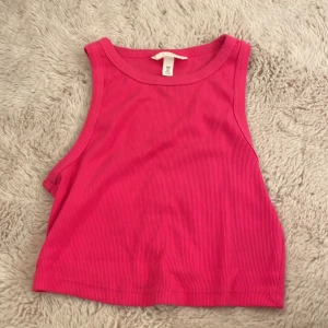 Rosa ribbad croppad linnetopp H&M - Säljer en snygg croppad linnetopp från H&M i stark rosa färg. Toppen är ribbad och ärmlös med rund halsringning, perfekt för varma dagar eller lager på lager. Enkel och stilren design som är lätt att matcha.