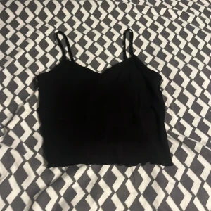 Svart croppad linnetopp från H&M - Svart croppad linnetopp från H&M med smala axelband och lätt v-ringad hals. Toppen har volangkant nertill som ger en söt touch. Perfekt till jeans eller kjol för en chill och stilren look.