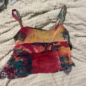 Färgglad linnetopp med volang - Unik linnetopp i tunnt material med färgstarkt mönster i rött, gult, grönt och blått. Toppen har smala axelband och en snygg volangdetalj framtill. Skit snygg topp för en utekväll. Den är kort och går över magen.