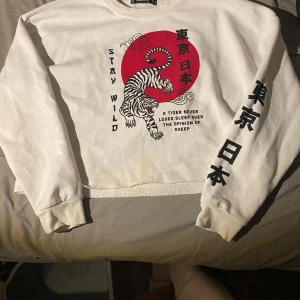 Vit sweatshirt med tigertryck och japansk text - Cool vit sweatshirt från Bershka med stort rött tryck och en tiger på framsidan. Japansk text på både bröstet och ena ärmen. Croppad modell med ribbade muddar och rund hals. Perfekt för dig som gillar streetstyle och unika prints. Rekommenderas för ungdom mellan 13-16 år