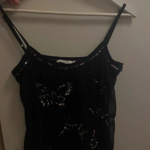 Svart paljettlinne från Vila - Supersnygg svart linnetopp från Vila med tunna axelband och glittriga paljettdetaljer i fjärilsmönster. Perfekt för dig som vill sticka ut med lite extra bling. Toppens material är lätt och genomskinligt, vilket ger en cool och trendig look.