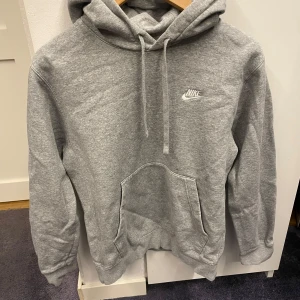 Grå hoodie från Nike med huva - Säljer en grå Nike hoodie i storlek XS med klassisk logga broderad på bröstet. Tröjan har huva med snörning, stor magficka och långa ärmar. Mjuk insida i fleece och relaxed passform, perfekt för chill eller träning. Fråga om du vill ha fler bilder!