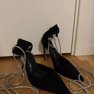Svarta klackskor med strassband och dragkedja - Snygga svarta klackskor med spetsig tå hög klack  (ca 10-12 cm) och glittrande strassband att snöra runt ankeln. Skorna har dragkedja bak för enkel påtagning och är tillverkade i mockaliknande material. Perfekta för dig som vill sticka ut med detaljer och glam. Skorna har använt bara en enda kväll sedan inget mer. Dock ser ni de smutsiga sulorna, men det är inget som påverkar användningen.