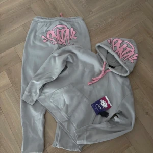 Grå tracksuit med rosa tryck från Syna world  - Säljer en grå hoodie från SNXH med fett rosa graffiti-tryck på huvan och matchande tjocka rosa snören. Hoodien har känguruficka och mjukt material, perfekt för chill dagar. Snygg streetwear-vibe och riktigt skön passform. Pris kan snackas vid snabba affärer 