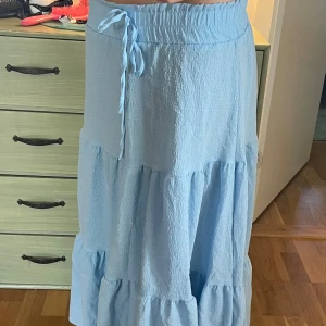 Ljusblå volangkjol med knytband - Säljer en ljusblå långkjol med volanger och knytband i midjan. Kjolen har en mjuk, krinklad struktur och elastisk midja. Vet vart den är från men storleken är XS och skulle passa nån som är 150cm-160cm.