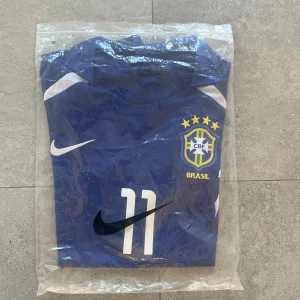 Blå Brasilien fotbollströja Nike Ronaldinho  - Snygg blå Brasilien fotbollströja från Nike med nummer 11 på bröstet med Ronaldinho på ryggen. Tröjan har vit Nike-logga, Brasilien-emblem med stjärnor och vita detaljer vid axlar och sidor. Tillverkad i Dri-Fit material som andas och har klassisk krage. Perfekt för dig som älskar fotboll och landslag. Pris kan diskuteras 
