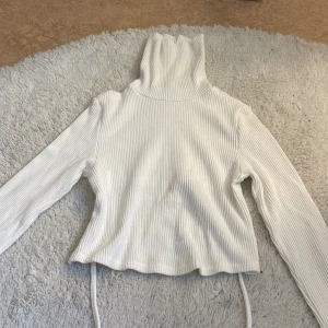 Vit ribbad Bershka tröja  med snörning - Säljer en vit ribbad Bershka tröja  med lång ärm och hög krage. Tröjan har snygga snörningsdetaljer i sidorna och är croppad med vågig kant nedtill. Perfekt för dig som gillar stilrena och trendiga plagg med lite edge.