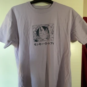 One Piece t-shirt från Pull&Bear - Ljuslila t-shirt från Pull&Bear med stort One Piece-tryck fram och bak. Motivet föreställer Luffy från mangan och text på japanska. Klassisk passform med rund hals och korta ärmar. Perfekt för animefans som vill sticka ut med stil.