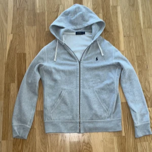 Grå hoodie från Polo Ralph Lauren storlek M - Snygg grå hoodie från Polo Ralph Lauren med klassisk logga broderad på bröstet. Tröjan har huva med snörning, dragkedja framtill och två fickor. Tillverkad i mjuk bomullsmix, perfekt för chill dagar. Passar dig som gillar stilren och enkel look. Dragkedjan har en nyckel ring som dragkedja för jag har råkat ta av den normala dragkedjan men jag ska sätta på en ny en vanlig. Storleken är M men skulle kunna passa S men vara lite för stor.