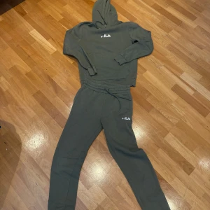 Gröna mjukisbyxor och tröja från Fila - Mörkgröna mjukisbyxor och tröja från Fila med broderad vit logga på benet och vid bröstet. Byxorna har resår och snörning i midjan för skön passform. Perfekta för chill eller träning, tillverkade i mjukt bomullsmaterial.