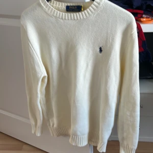 Vit stickad tröja från Polo Ralph Lauren - Säljer en klassisk vit stickad tröja från Polo Ralph Lauren med rund hals och diskret broderad logga på bröstet. Tröjan har långa ärmar och ribbade muddar vid ärmslut och nederkant. Perfekt för dig som gillar stilrena och tidlösa plagg.