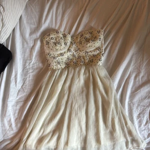 Champagnefärgad pärlklänning från Bik Bok - Supersöt kortklänning från Bik Bok i champagnefärg med pärlbroderad bustier och plisserad kjol. Klänningen är axelbandslös och har dragkedja i ryggen. Perfekt för student eller sommarens speciella tillfällen. Materialet känns lätt och festligt, med pärlor i olika färger på överdelen.