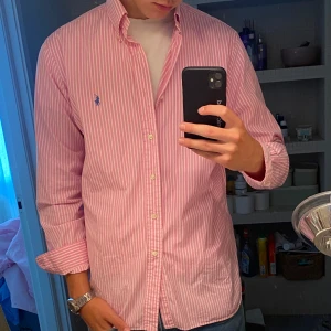 Rosa randig skjorta från Polo Ralph Lauren - Snygg rosa och vit randig skjorta från Polo Ralph Lauren med klassisk button down-krage och broderad logga på bröstet. Skjortan är långärmad och har knappar hela vägen framtill. Perfekt för dig som gillar en preppy och fräsch stil.