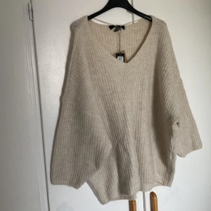Oversized beige stickad tröja V-ringad - Supermysig oversized stickad tröja från VERO MODA i beige ton. Tröjan har en djup v-ringning, ribbad struktur och långa, lösaå ärmar. Perfekt för lager-på-lager och chill dagar. Passar dig som gillar en relaxed och trendig look.