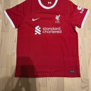 Liverpool FC röd Nike matchtröja - Liverpool FCs officiella matchtröja i rött med vita detaljer från Nike. Tröjan har korta ärmar, rund halsringning och klubbens logga samt sponsortryck på bröstet. Tillverkad i lätt och ventilerande material som passar perfekt för fotboll eller träning.