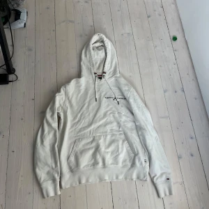 Vit hoodie från Tommy Hilfiger  - Snygg vit hoodie från Tommy Hilfiger i storlek M. Klassisk design med broderad logga och text på bröstet, känguruficka och huva med snörning. Ribbade muddar och mjukt bomullsmaterial gör den riktigt skön att bära.