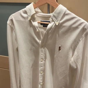 Vit skjorta från Ralph Lauren - Klassisk vit skjorta från Ralph Lauren med broderad logga på bröstet. Skjortan har button-down krage, långa ärmar och knäppning framtill. Tillverkad i mjuk bomull som känns skön mot huden. Perfekt för en clean och stilren look.