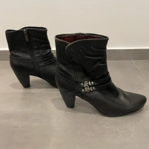 Svarta stövletter från Tamaris - Svarta stövletter i skinn från Tamaris med snygga detaljer i form av spännen och remmar runt ankeln. Skorna har dragkedja på sidan, rund tå och stabil klack. Insidan är fodrad i vinrött tyg för extra komfort. Perfekta för höst och vinter.