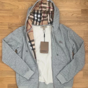 Grå hoodiejacka från Burberry - Grå hoodiejacka från Burberry med klassiskt rutigt foder i huvan. Jackan har dragkedja, snörning i huvan och ribbade muddar. Perfekt för dig som vill ha en stilren och lyxig look med ikoniska Burberry-detaljer.