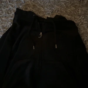 Svart hoodiejacka från Juicy Couture - Svart hoodiejacka från Juicy Couture med dragkedja framtill och snörning i huvan. Klassisk streetstil med kängurufickor och mjukt material som känns skönt mot huden. Perfekt för en chill och avslappnad look.