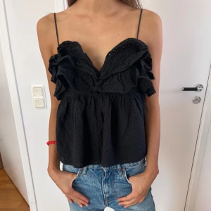 Svart volangig blus med tunna axelband - Supersöt svart blus med tunna axelband, öppen rygg och volangdetaljer framtill. Toppen har broderade hålmönster. Använde en gång 