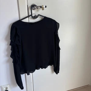 Svart blus med volangärmar - Säljer en svart blus med långa ärmar och snygga volangdetaljer längs ärmarna. Blusen har en lätt och luftig känsla, perfekt för dig som gillar stilrena plagg med en twist. Passar till både jeans och kjol för en trendig look.