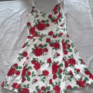 Vit kortklänning med röda rosor - Superfin vit kortklänning med röda rosor och gröna blad. Klänningen har ärmlös design, rundad hals och ett lätt utställt kjolparti som ger en skön och ungdomlig vibe. Perfekt för varma dagar och somriga tillfällen.