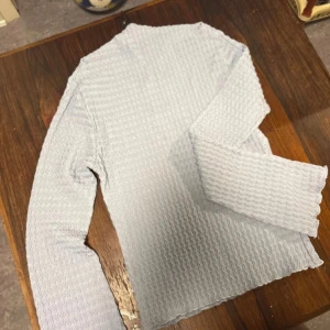 Ljusblå ribbad långärmad tröja - Säljer en ljusblå långärmad tröja med ribbad struktur och vågigt mönster. Tröjan har en enkel, rak passform och är tillverkad i ett mjukt material som känns skönt mot huden. Perfekt för dig som gillar stilrena och bekväma plagg.