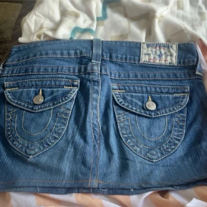 True Religion jeanskjol med fickor - Säljer en blå jeanskjol från True Religion med snygga bakfickor och detaljerade sömmar. Kjolen har knappstängning fram, bälteshällor och klassisk jeanslook. Perfekt för dig som gillar streetstyle och vill ha en ikonisk denimkjol i garderoben.