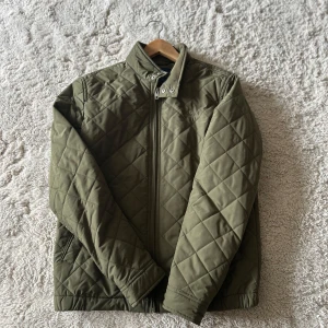 Olivgrön quiltad jacka från GANT - Snygg olivgrön quiltad jacka från GANT med dragkedja och hög krage. Jackan har ett stilrent rutmönster, två fickor och diskret logga på bröstet. Perfekt för dig som vill ha en clean look under höst och vår. Priset går att diskutera 