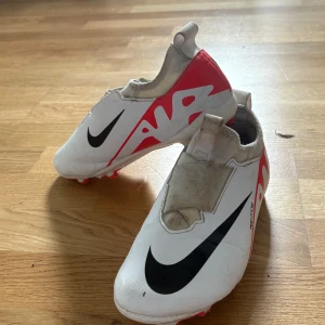 Nike Air Zoom fotbollsskor vit/röd - Snygga Nike Air Zoom fotbollsskor i vitt med röda och svarta detaljer. Skorna har en stilren design med dold snörning och dragflik bak. Ovandelen är i syntetmaterial och sulan har röda dobbar för bra grepp på planen.