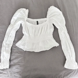 Vit blus med puffärm från H&M Divided - Supersöt vit blus från H&M Divided med puffiga långa ärmar och smockad midja. Den har fyrkantig ringning, volang nertill och elastiska detaljer som ger en snygg passform. Perfekt för dig som gillar romantisk och trendig stil.
