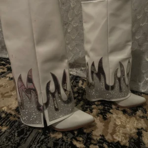 Vita cowboy boots med silver flames - Unika vita cowboy boots med spetsig tå och coola silverglittriga flames längs skaftet. Boots i syntetmaterial med metallicdetaljer och platt sula. Perfekta för dig som vill sticka ut med en statement-look.