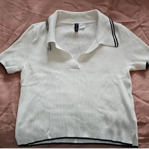 Vit ribbad pikétröja från H&M Divided - Vit ribbad pikétröja från H&M Divided med svarta detaljer på krage, ärmslut och nederkant. Tröjan har korta ärmar, klassisk pikékrage och är tillverkad i mjuk viskosblandning. Perfekt för en clean och sportig look.