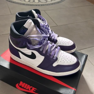 Nike Air Jordan 1 Court Purple High - Nike Air Jordan 1 High i färgen Court Purple med vit bas, svarta detaljer och lila overlays. Klassisk svart swoosh, perforerad tå och lila skosnören. Ovandel i skinn och hög siluett som ger en ikonisk streetwear-look. Perfekta för dig som vill sticka ut med sneakers. Köpta från soleplus, använda varsamt med crease skydd och svarta snören skickas med.