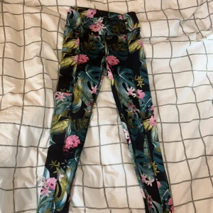 tränings leggins  - Snygga leggings från SOC med tropiskt blommönster i rosa, grönt och blått på svart botten. Tight passform och hög midja, perfekta för träning eller chill. Materialet är stretchigt och andas, så du håller dig bekväm hela dagen. Stl 134/140