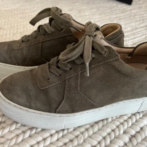 Olivgröna sneakers från Axel Arigato - Snygga olivgröna sneakers från Axel Arigato i mocka med vita, platta sulor och klassisk snörning. Skorna har rund tå och diskret logga i guld på sidan. Perfekta för dig som gillar stilrena och trendiga sneakers med premiumkänsla.